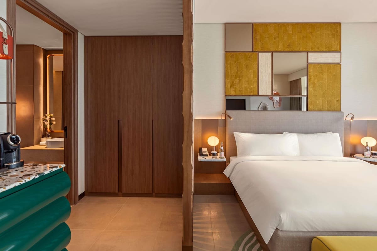 A hotel room with a wooden wardrobe, a bed with white bedding, a bedside table with a lamp, and a mirror above the bed.