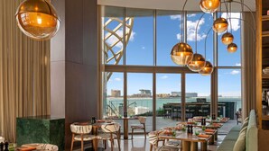 A restaurant with multiple tables and chairs, large windows with a view of the city, and hanging pendant lights. - So/ Ras Al Khaimah Hotel & Resort (Ras Al Khaimah)