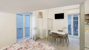 Un dormitorio con cama, escritorio, silla, balcón y televisor montado en la pared.