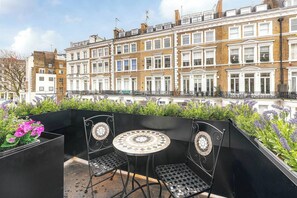 Eine Dachterrasse mit einem runden Tisch und zwei Stühlen, umgeben von einem schwarzen Metallzaun und Pflanzkübeln mit violetten Blumen.