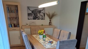 Ein Essbereich mit Holztisch, grau gepolsterten Stühlen, einer Hängeleuchte sowie eine Küchenzeile mit Schränken und einer Vase.