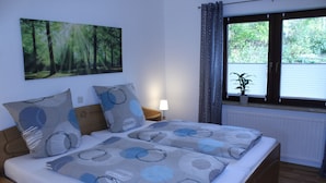Ein Doppelbett mit gemustertem Bettbezug, ein Nachttisch mit Lampe, ein Fenster mit Vorhängen und ein Wandbild von einem Wald.