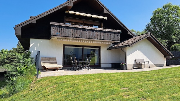 Ein zweistöckiges Haus mit Balkon, einer Terrasse mit Stühlen und einem Rasen.