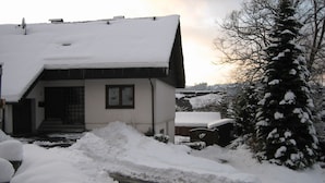 Ein Haus mit geneigtem, schneebedecktem Dach, einem sichtbaren Fenster und einem mit Schnee bedeckten Baum im Vordergrund.