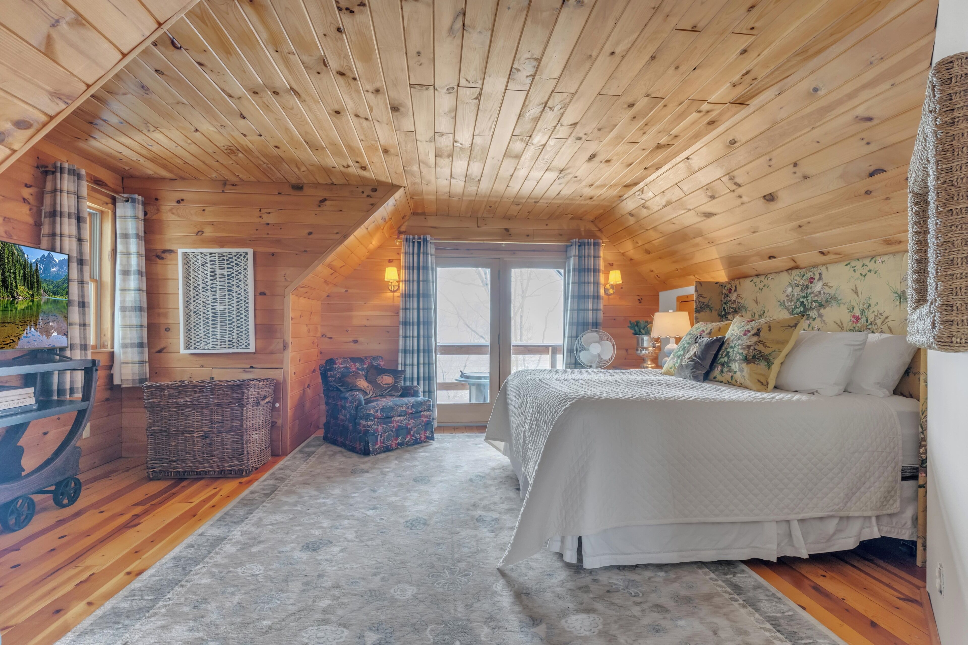 A cozy attic bedroom with a wooden ceiling, a bed with a white duvet, a wicker basket, a blue patterned chair, and a small table with a lamp.