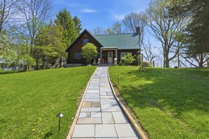 Une maison en bois avec un toit vert, un chemin pavé menant à l’entrée et une cour gazonnée avec des arbres.