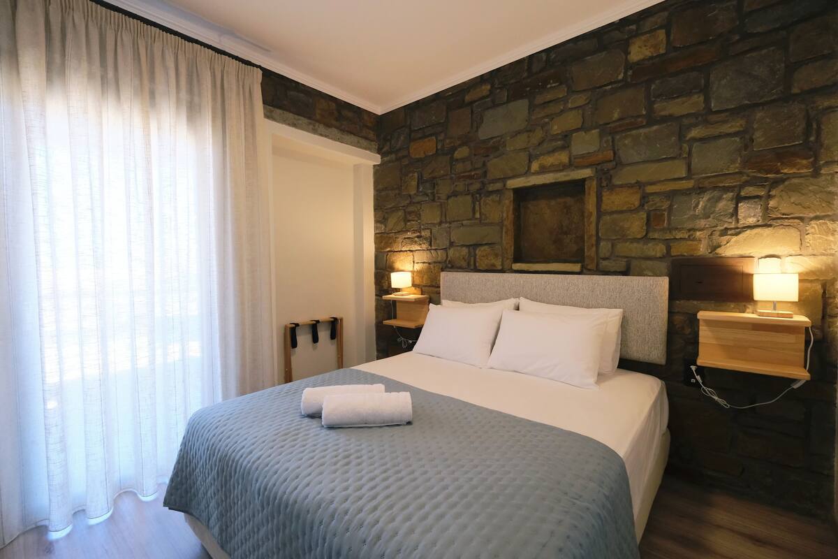 A hotel room with a stone wall, a bed with white bedding, two bedside lamps, and a window with sheer curtains.