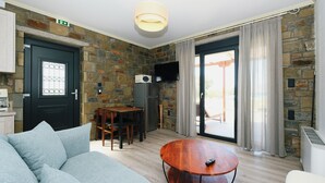 A modern living room with a stone wall, a black door, a dining area with a table and chairs, a round wooden coffee table, and a television mounted on the wall. - Amari stone apartments (Lemnos)