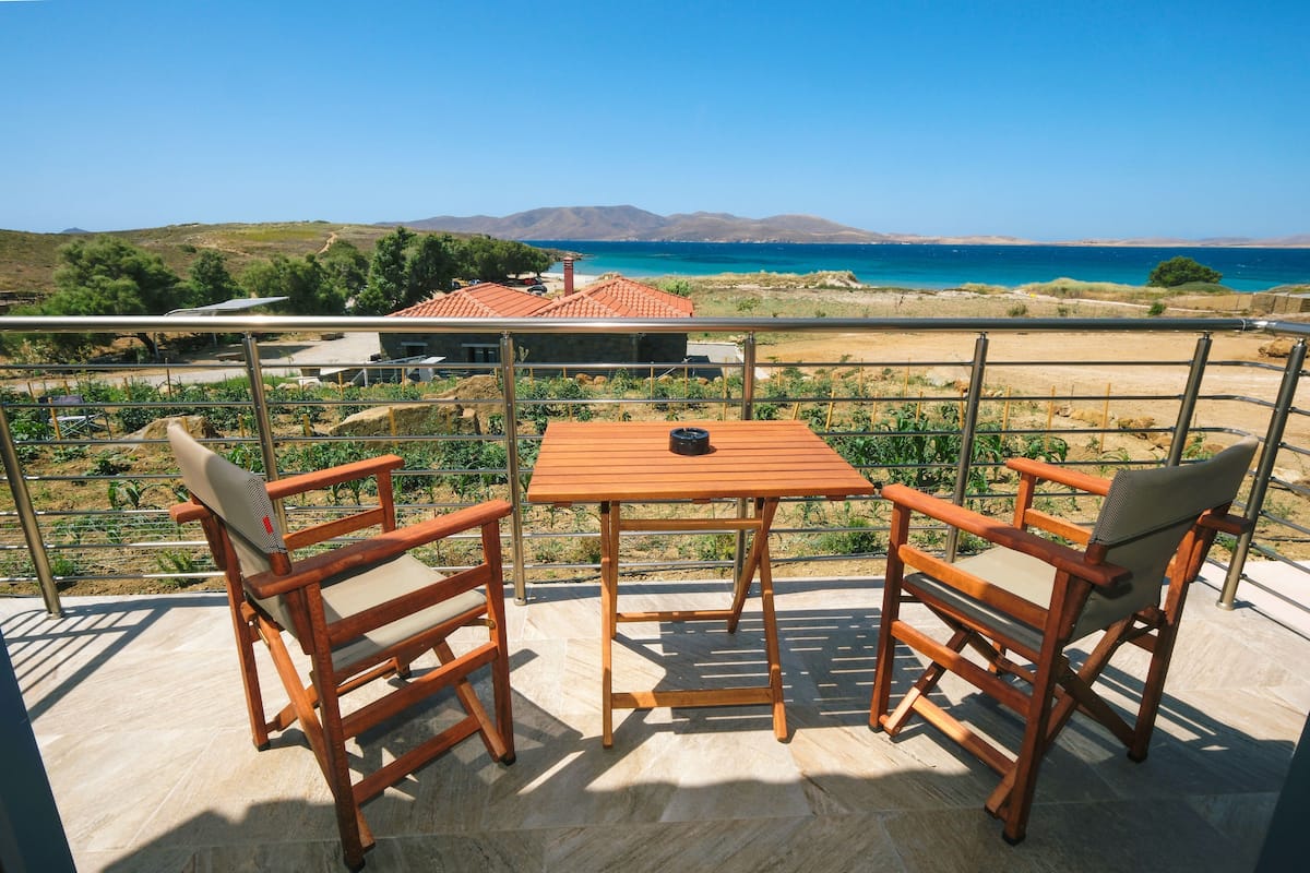 A balcony with two wooden chairs and a table, overlooking a coastal landscape with mountains in the distance.