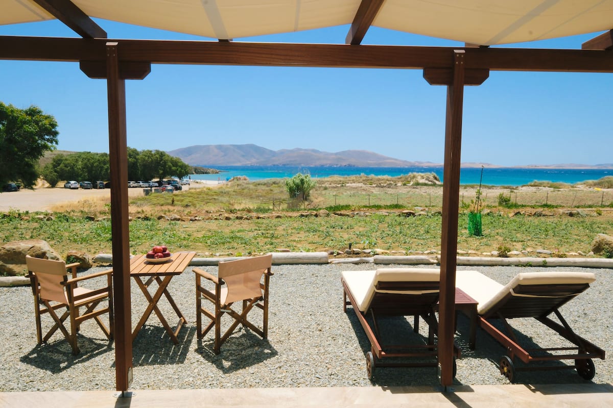 A covered outdoor seating area with a table and chairs, lounge chairs, and a view of the sea and mountains.