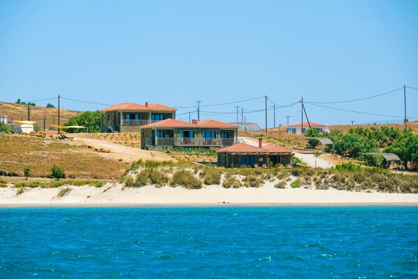Una propiedad costera con varias construcciones, incluyendo una casa principal y estructuras más pequeñas, ubicada en una playa de arena con aguas azules y transparentes al frente.