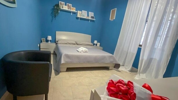 Een slaapkamer met een bed, een stoel, een nachtkastje en een wandplank met foto's.