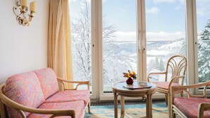 A cozy room with a sofa, two chairs, a small table with a vase, and a large window with a view of snow-covered trees and a distant landscape. - Bergvital Hotel Todtnau (Todtnau)