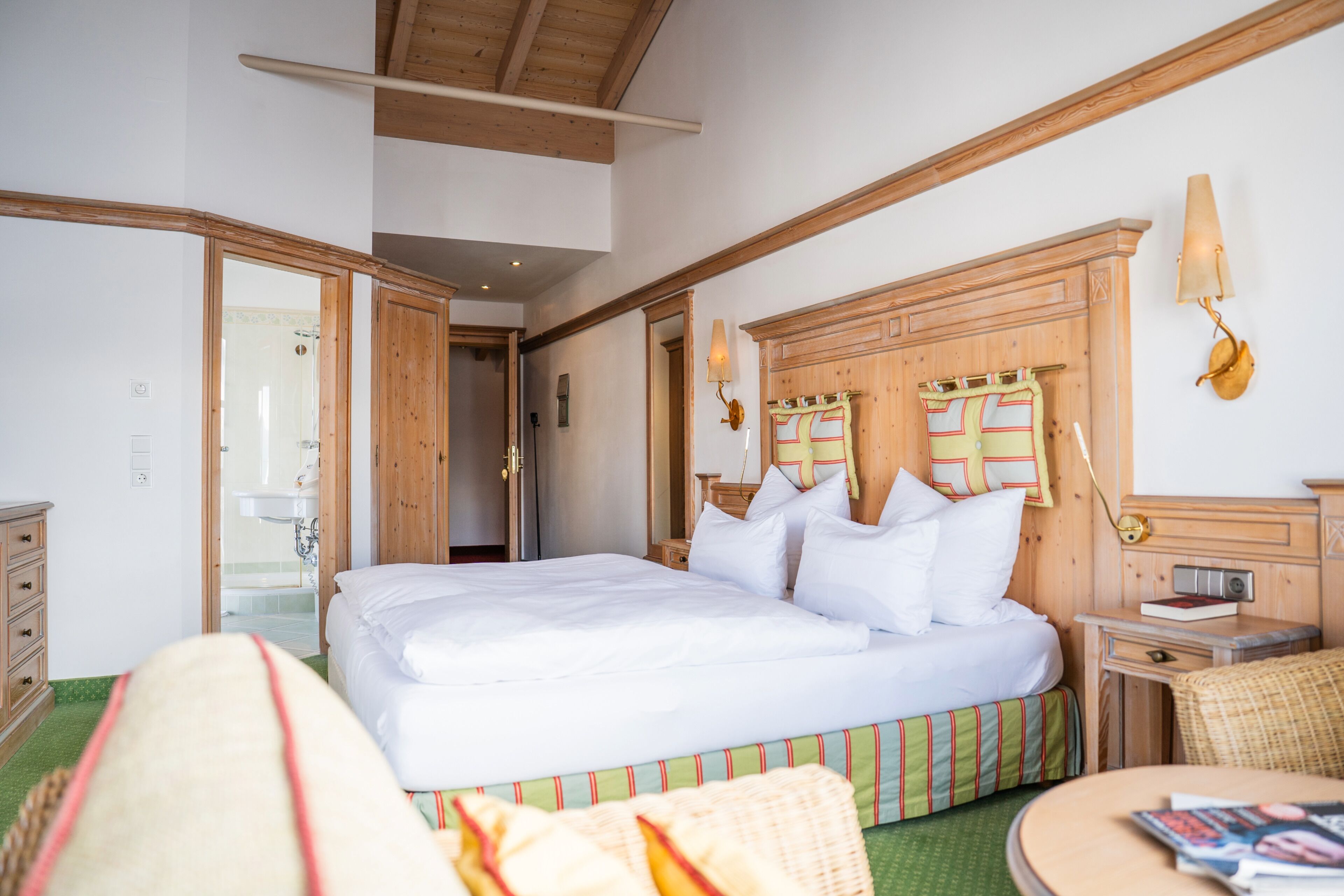 Habitación de hotel con una cama grande, cabecera de madera, una mesita de noche con lámpara y vistas al baño a través de una puerta abierta.