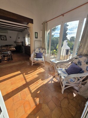 A spacious living area with a large window overlooking a garden, furnished with wicker chairs and a wooden table. - Nathalia House (Panorama)