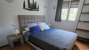 Un dormitorio con cama, mesita de noche, lámpara, ventana con cortinas y repisa en la pared.
