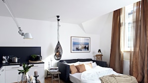 Ein modernes Hotelzimmer mit Bett, Esstisch mit Stühlen, Küchenzeile und großem Fenster mit Vorhängen.