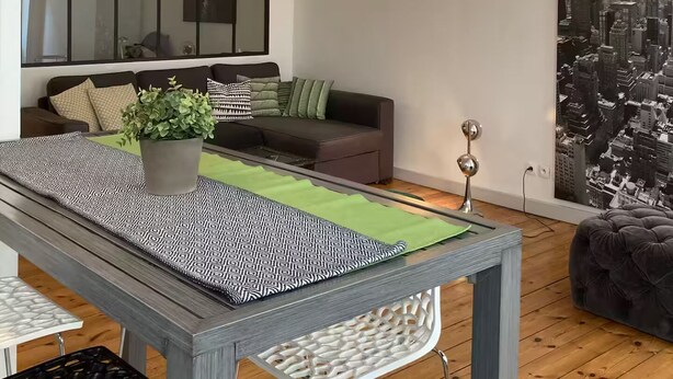 A modern living room with a grey sofa, a dining table with patterned table runners, a potted plant, and a cityscape wall art. - Aux Pieds des Tours (La Rochelle)