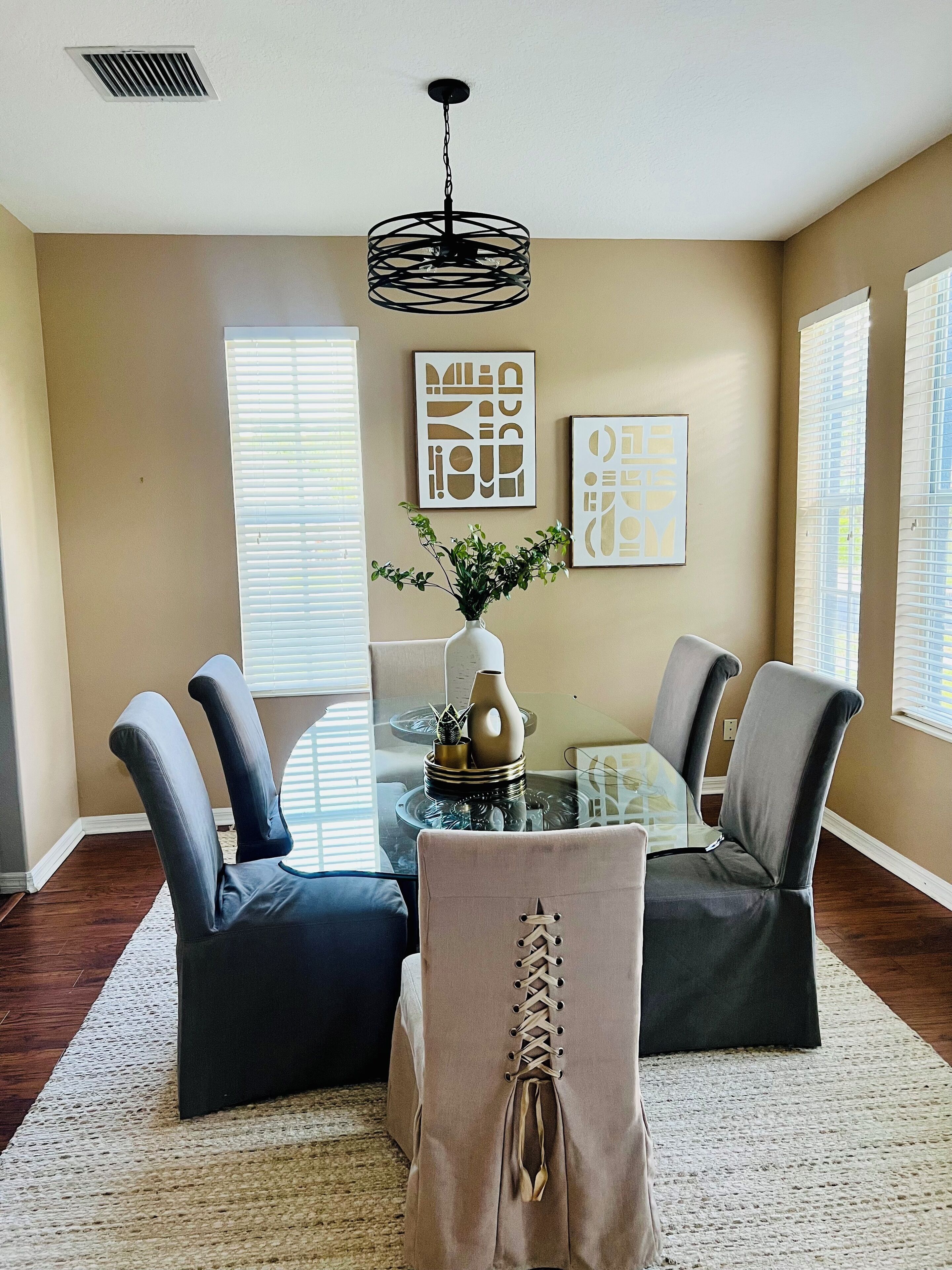A dining area with a glass table, two grey chairs, a vase with greenery, and abstract wall art.