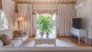Una habitación con sofá, una mesa pequeña con un jarrón de flores, televisor y ventana con cortinas.