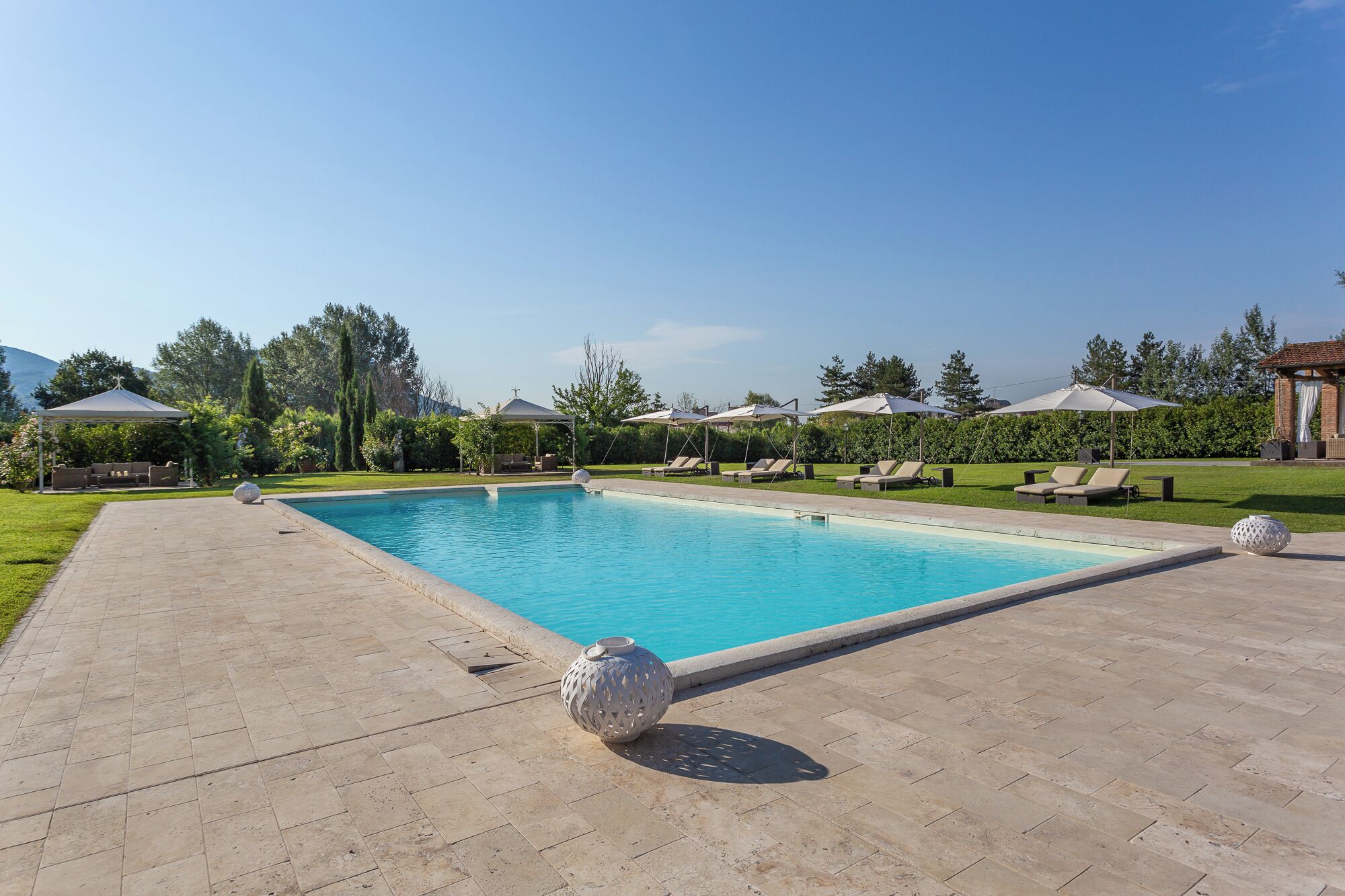 Une piscine rectangulaire, bordée d’une terrasse carrelée, entourée de chaises longues et de parasols, avec un belvédère et de la verdure en arrière-plan.