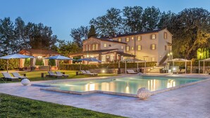 Un área junto a la piscina con sillones de descanso y sombrillas, piscina y un edificio grande al fondo.