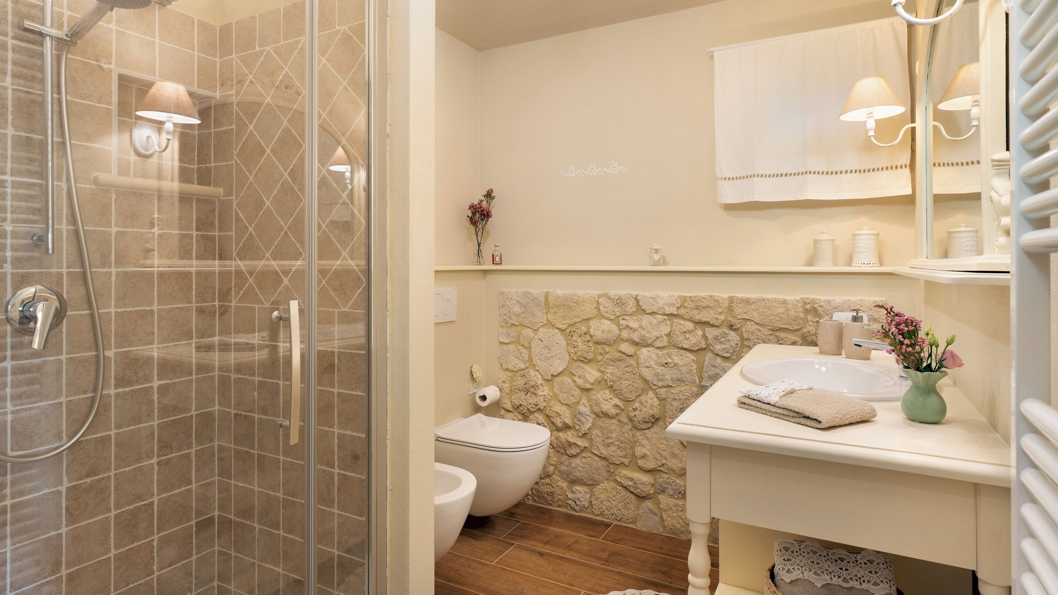A bathroom with a glass-enclosed shower, a toilet, a sink with a vase of flowers, and a mirror with a towel hanging on the wall.