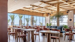 A spacious restaurant with wooden tables and chairs, large windows with a view of palm trees, and a ceiling with a wooden pattern. - Kakkos Terra Blu (Ierapetra)