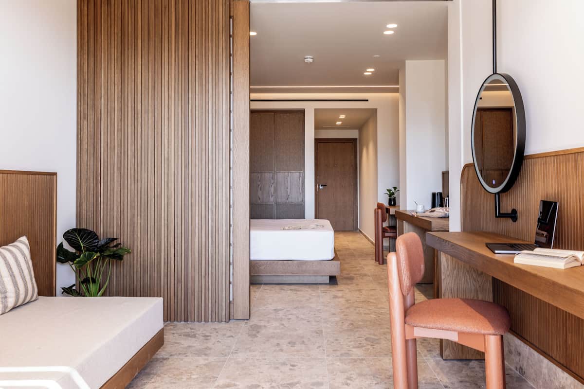 A modern hotel room with a bed, a desk, a chair, a mirror, and a plant.