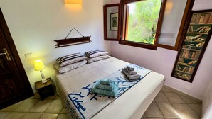 Ein ordentlich bezogenes Bett mit gestreiften Kissen, eine Nachttischlampe, ein an der Wand befestigtes Regal und ein Fenster mit Blick auf Grünflächen.