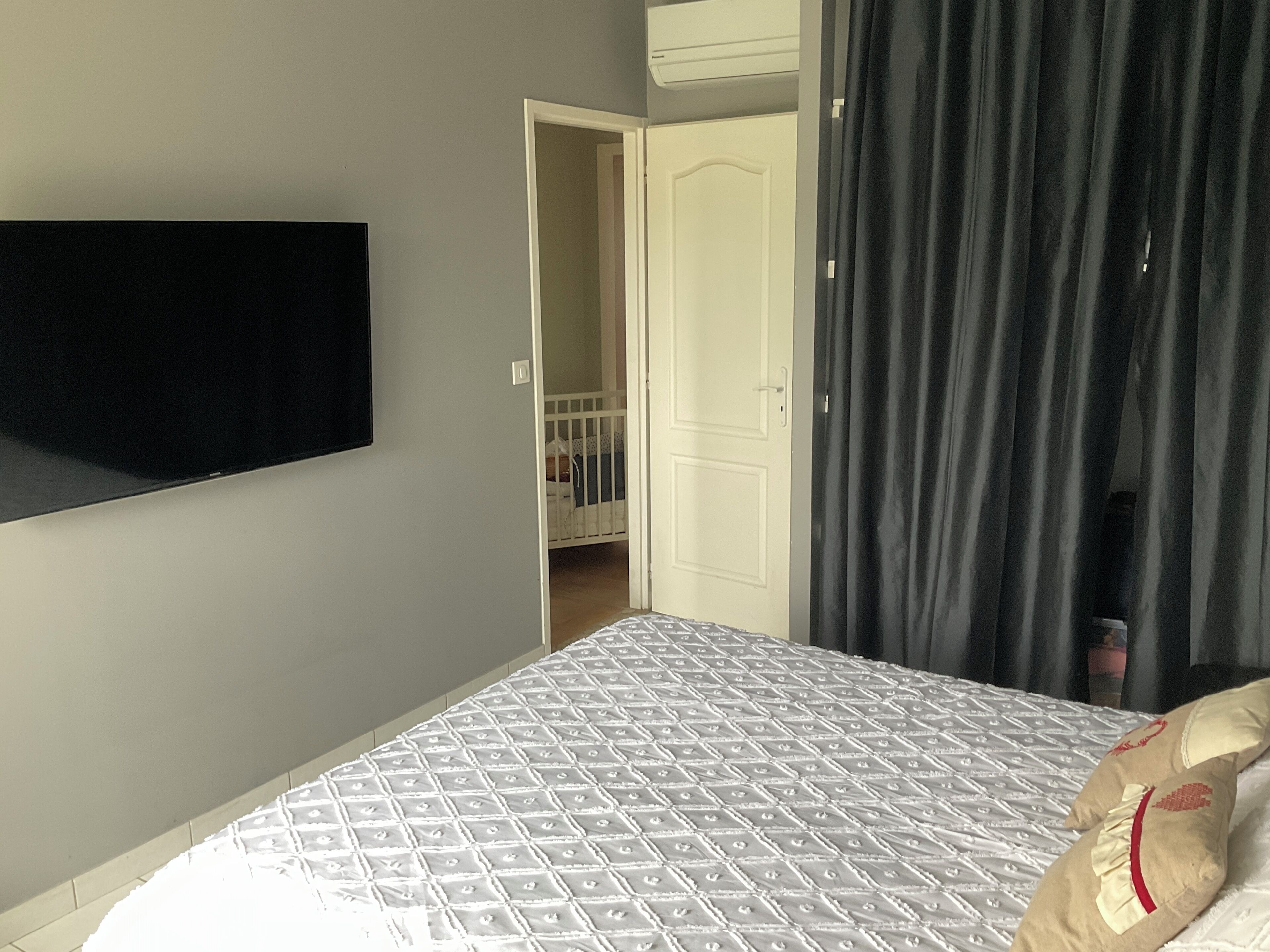 A hotel room with a bed, a TV mounted on the wall, a door leading to a balcony, and a window with curtains.