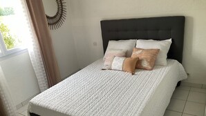 Un lit bien fait, avec une couette blanche et plusieurs oreillers, une tête de lit sombre et une vue sur l’extérieur par une fenêtre ornée de rideaux.