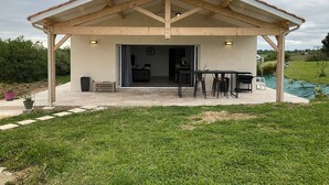 Un espace extérieur abrité, équipé d’une table et de chaises, d’un petit banc et d’une porte donnant sur un espace intérieur.