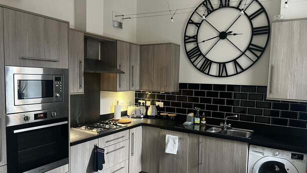 Une cuisine moderne avec des meubles en bois, des appareils en acier inoxydable, une grande horloge murale et un dosseret de carrelage noir et blanc.