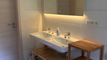 A modern bathroom with a double sink, a mirror, and a wooden shelf.