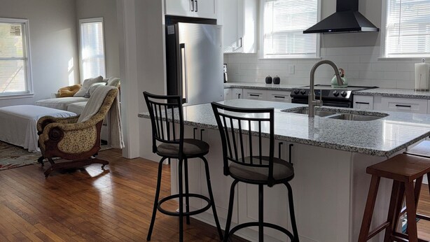 A modern kitchen with white cabinetry, a black range hood, and a granite countertop. There are two bar stools and a wooden chair. - Beautiful 3-bedroom house with AC in Birmingham's Entertainment District. (Birmingham)