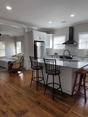 Una cocina moderna con armarios blancos, una campana extractora negra y una encimera de granito. Hay dos taburetes de bar y una silla de madera.