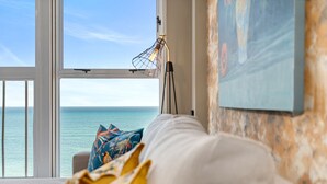 Ett rum med utsikt över havet, en vit soffa med färgglada kuddar och en lampa med en lampskärm av metalltråd.