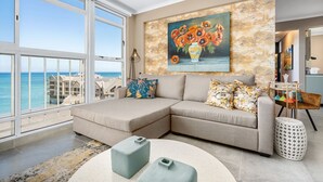 Ett modernt vardagsrum med ett stort fönster med utsikt över havet, en beige hörnsoffa och en tavla med blommor på väggen.