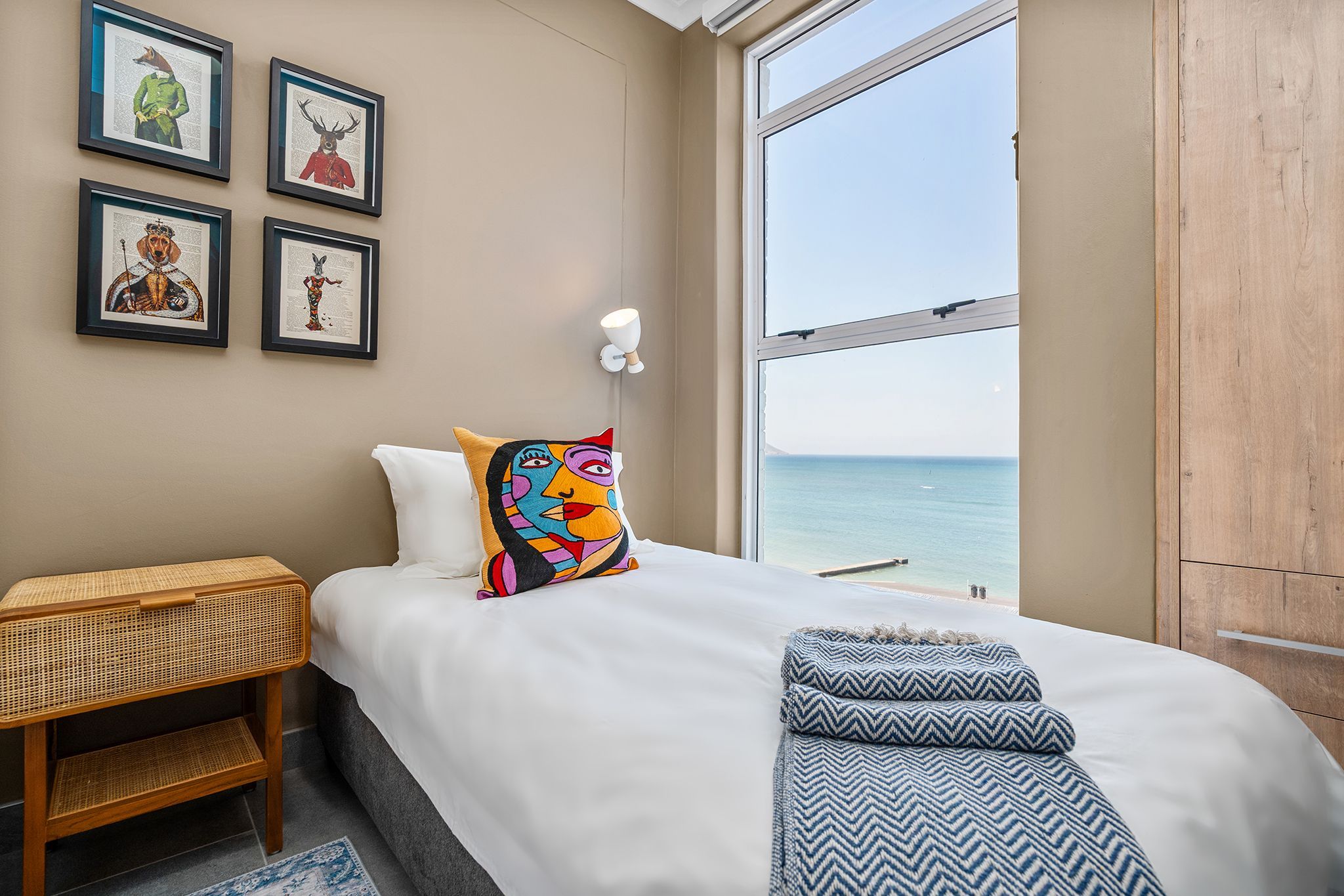 A bedroom with a large window, a bed with a colorful pillow, a small side table, and four framed artworks on the wall.