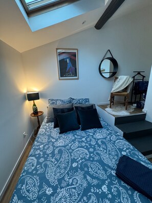 A bedroom with a bed, a nightstand, a lamp, a framed poster, a mirror, and a chair with a blanket. - Masion de ville Champenoise (Épernay)