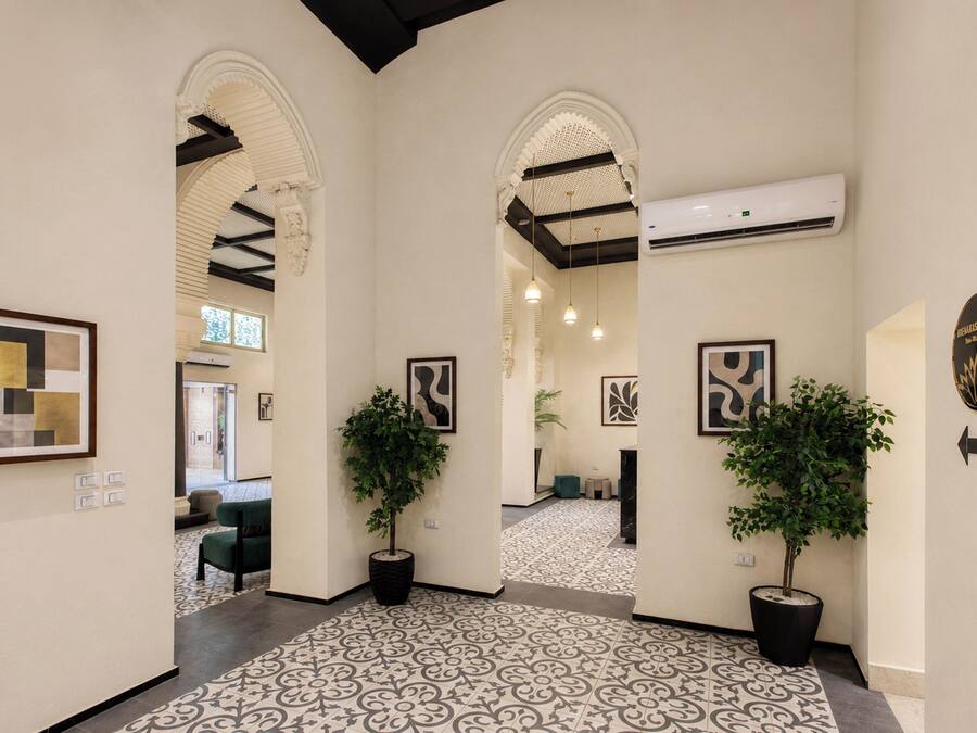 A hallway with patterned flooring, two potted plants, framed artwork, and an air conditioning unit.