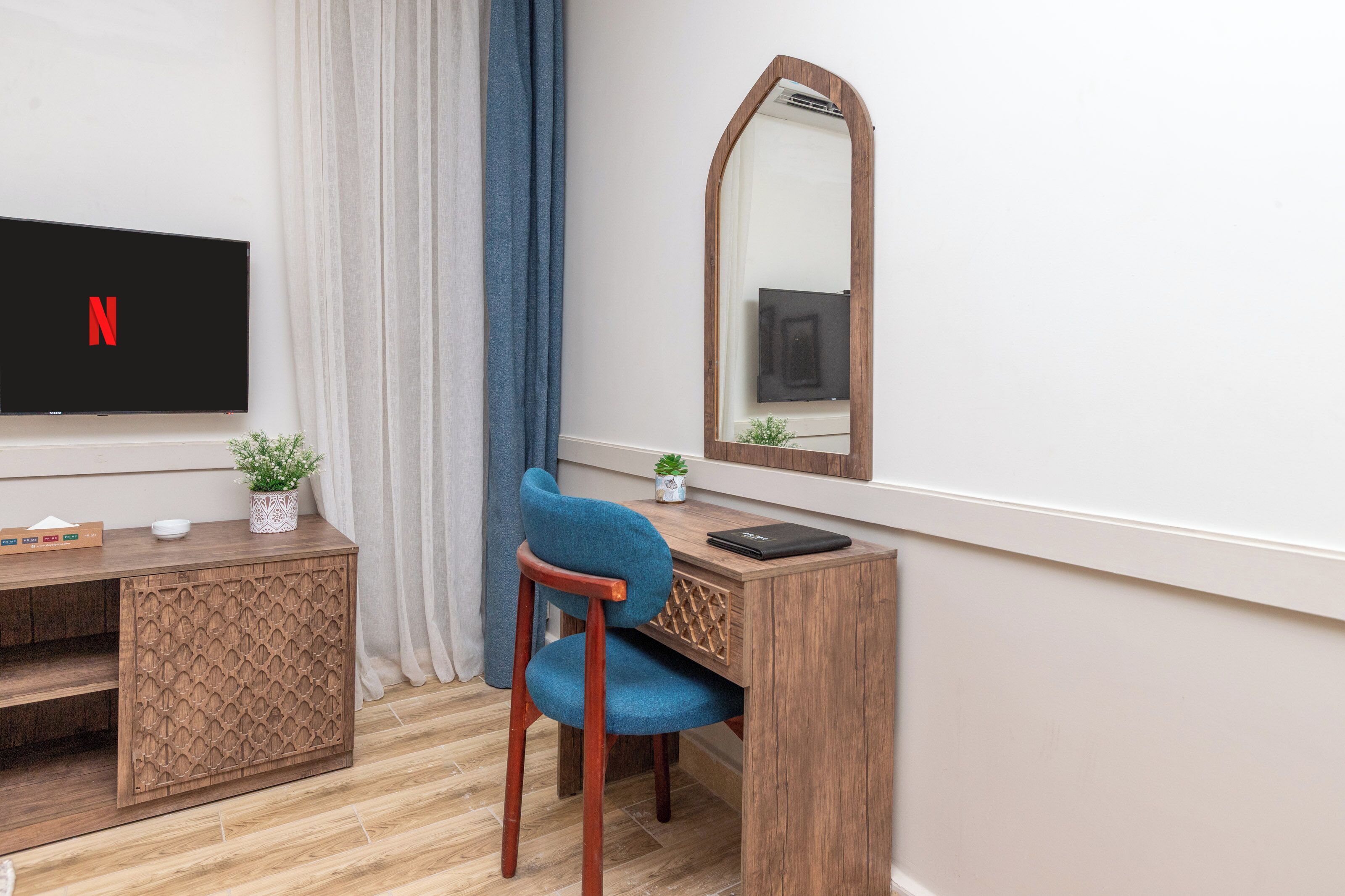 Una habitación con una televisión mostrando el logo de Netflix, un escritorio de madera con una computadora portátil, una silla, una planta pequeña y un espejo.