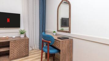 Una habitación con una televisión mostrando el logo de Netflix, un escritorio de madera con una computadora portátil, una silla, una planta pequeña y un espejo.