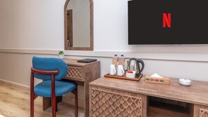 Une chambre d’hôtel avec un bureau en bois, une chaise bleue, un miroir, un téléviseur affichant le logo de Netflix, et un plateau avec une cafetière et des mouchoirs.