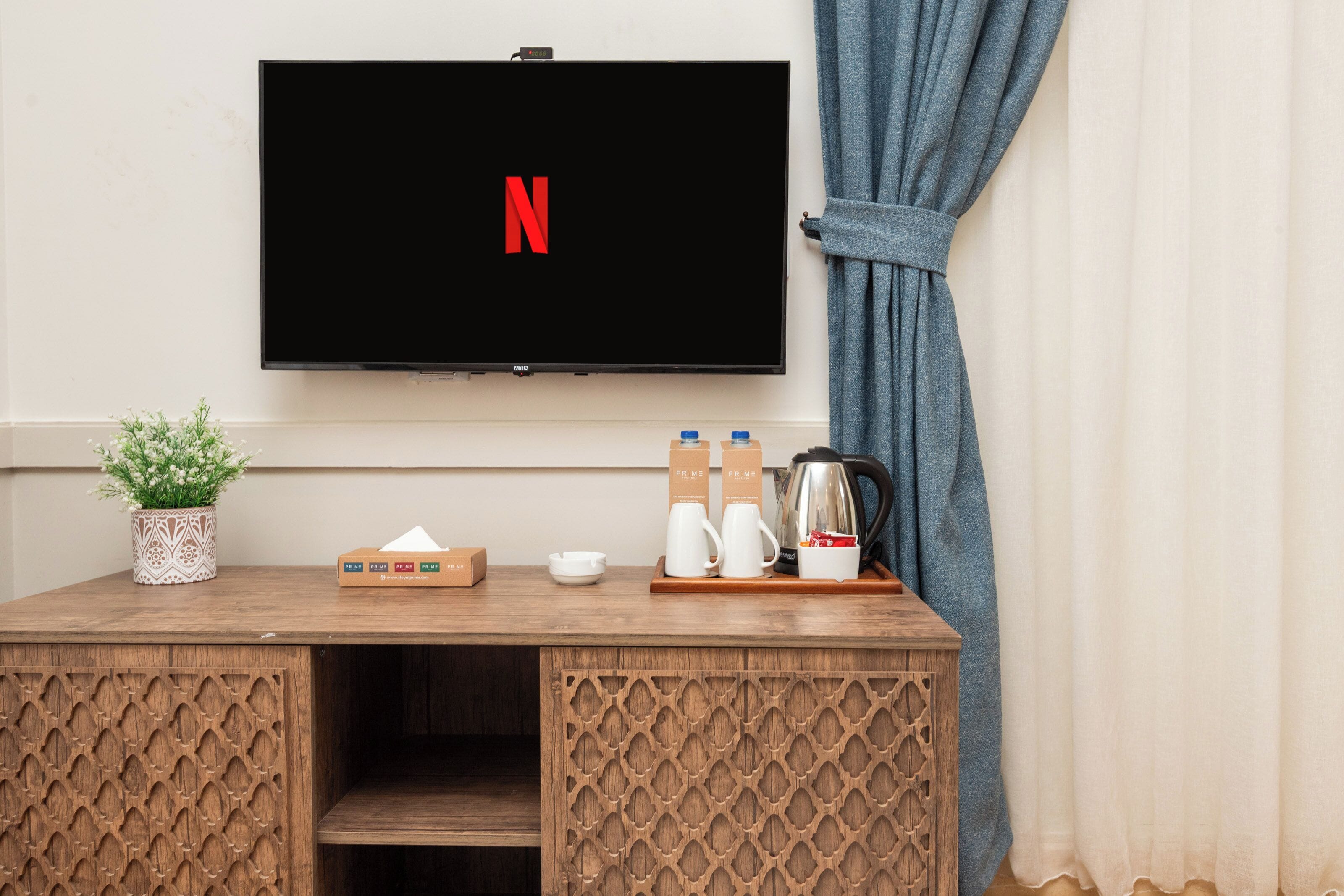 Et vægmonteret fladskærms-TV med Netflix-logo, et træbord med en potteplante, en tissueskasse, en kaffekande og to kopper.
