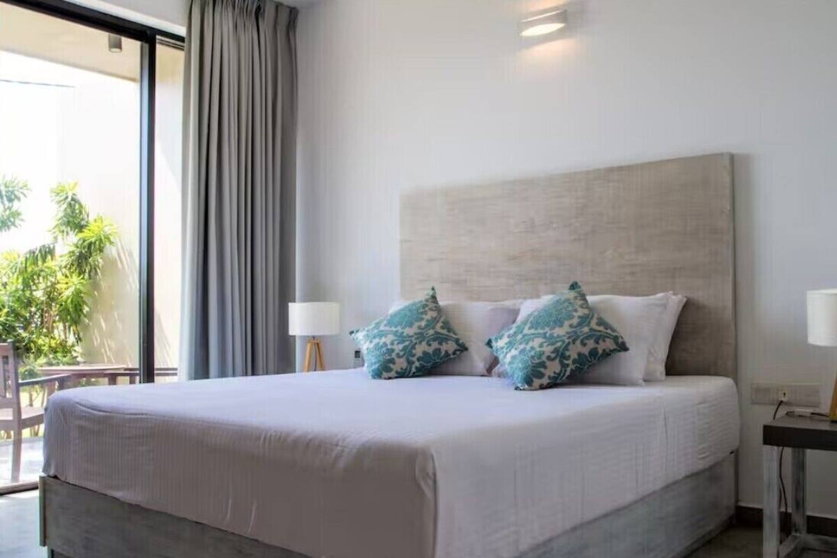 A hotel room with a large bed, two pillows, a nightstand, and a view of greenery outside.