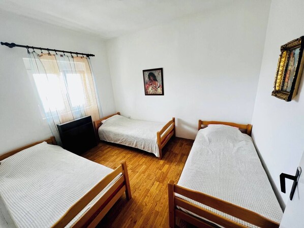 A room with three wooden beds, a black chair, a window with sheer curtains, and a framed picture on the wall.