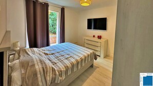 Una camera da letto con un letto, una TV montata a parete, un comodino con due candele rosse e una finestra con tende.