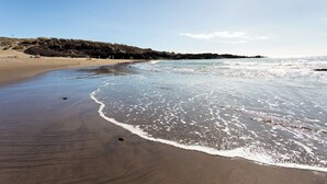 Een zandstrand met zachte golven, een heldere lucht en rotsformaties in de verte.